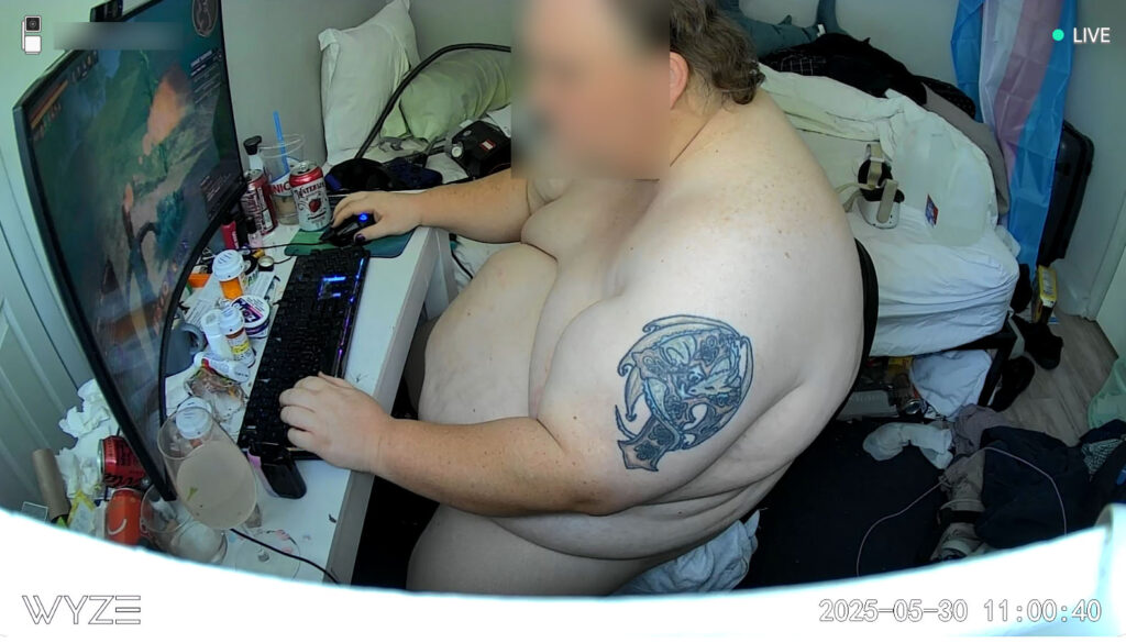 Overweight man caught shirtless on a surveillance cam in a cluttered room, surrounded by junk and medication — a raw display of humiliation under full digital control.